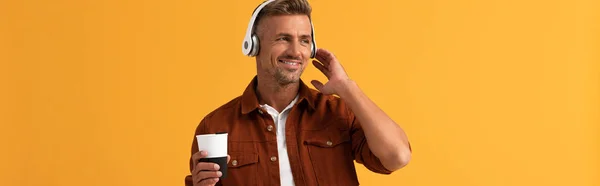 Panoramic shot of happy man listening music and holding coffee to go isolated on orange — Stock Photo