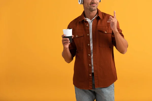 Abgeschnittene Ansicht eines glücklichen Mannes, der Musik hört und mit dem Finger zeigt, während er Kaffee to go isoliert auf orange hält — Stockfoto