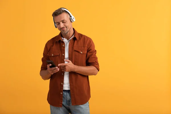 Hombre feliz escuchando música y señalando con el dedo al teléfono inteligente aislado en naranja - foto de stock