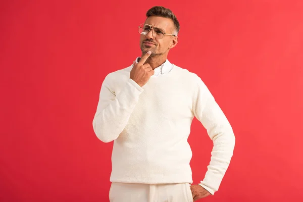 Pensive man in glasses standing with hand in pocket and touching face isolated on red — Stock Photo