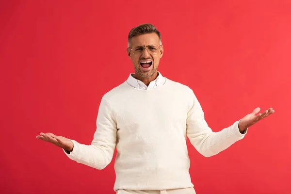 Hombre enojado en gafas gritando mientras está de pie con las manos extendidas aisladas en rojo - foto de stock