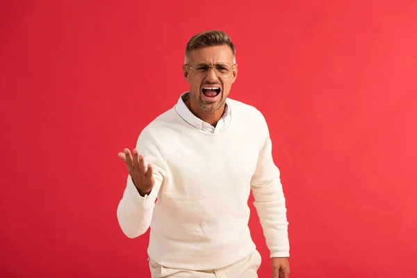 Hombre enojado en gafas gritando mientras está de pie y haciendo gestos aislados en rojo - foto de stock