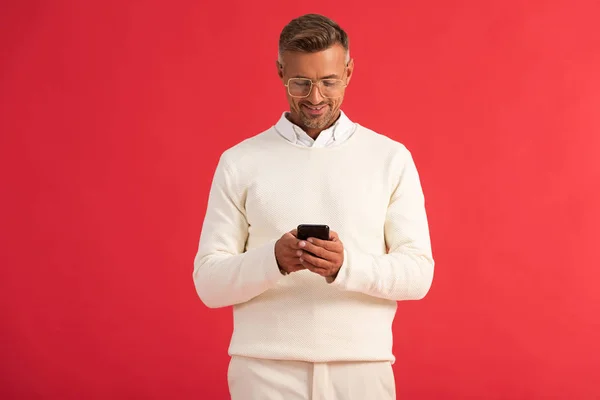 Hombre feliz en gafas usando teléfono inteligente aislado en rojo - foto de stock