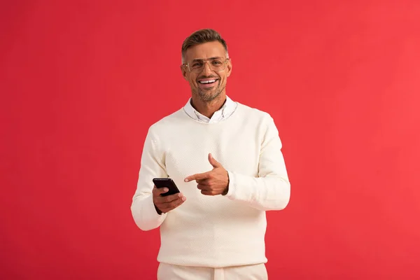 Hombre feliz en gafas apuntando con el dedo al teléfono inteligente aislado en rojo - foto de stock