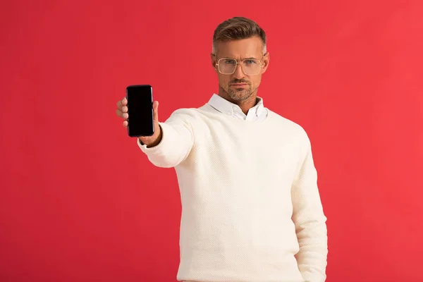 Bel homme dans des lunettes tenant smartphone avec écran blanc isolé sur rouge — Photo de stock