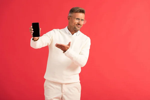 Uomo scontento in occhiali che punta con mano a smartphone con schermo vuoto isolato sul rosso — Stock Photo