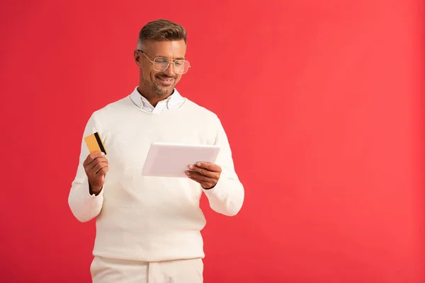 Hombre alegre sosteniendo tableta digital y tarjeta de crédito aislado en rojo - foto de stock