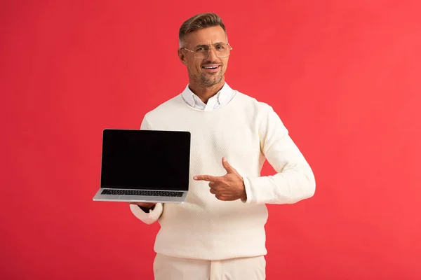 Homem feliz em óculos apontando com o dedo para laptop com tela em branco isolado no vermelho — Fotografia de Stock