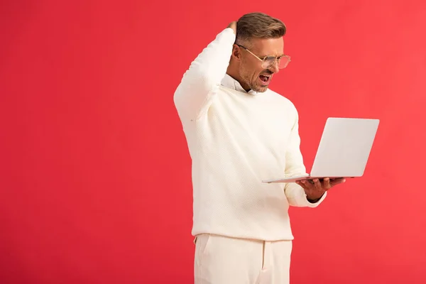 Незадоволений чоловік в окулярах тримає ноутбук ізольовано на червоному — стокове фото