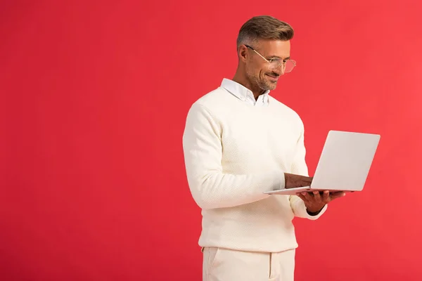 Uomo felice ed elegante in occhiali che tiene il computer portatile isolato su rosso — Foto stock