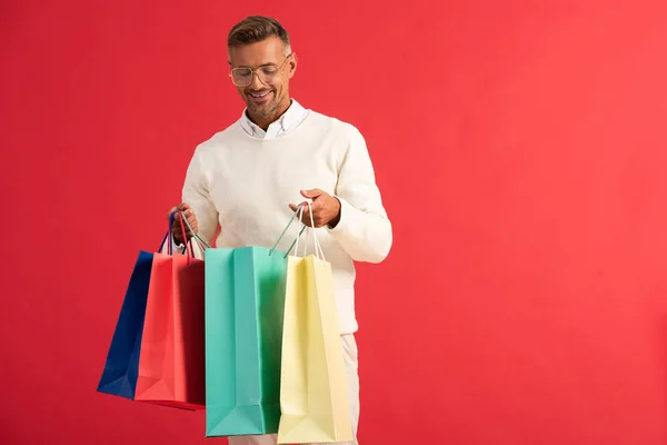 Hombre alegre en gafas sosteniendo coloridas bolsas de compras aisladas en rojo - foto de stock