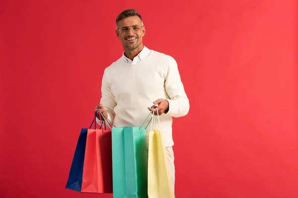 Happy man in glasses holding colorful shopping bags isolated on red — Stock Photo
