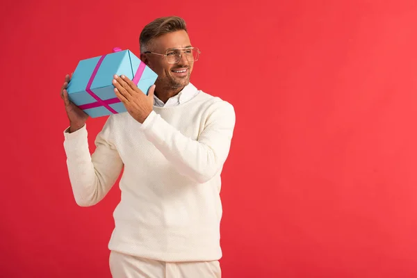 Happy man in glasses holding gift box isolated on red — Stock Photo