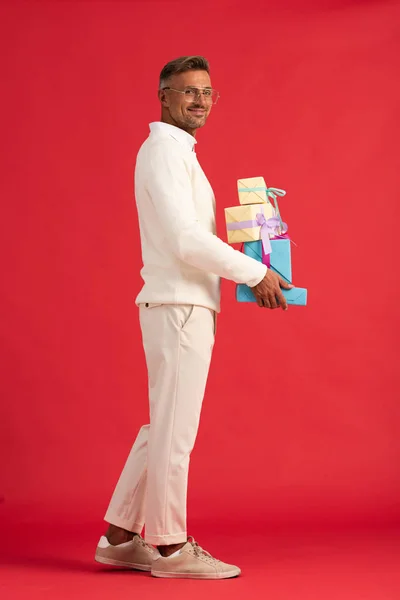 Cheerful man in glasses holding gift boxes while standing on red — Stock Photo