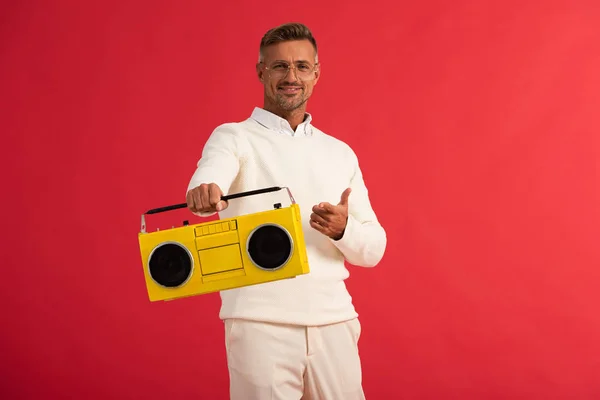 Hombre feliz sosteniendo boombox retro y señalando con el dedo aislado en rojo - foto de stock