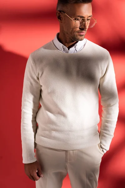 Homme élégant dans des lunettes debout avec la main dans la poche sur rouge avec des ombres — Photo de stock