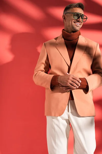 Uomo alla moda in occhiali da sole e blazer in piedi sul rosso con ombre — Foto stock