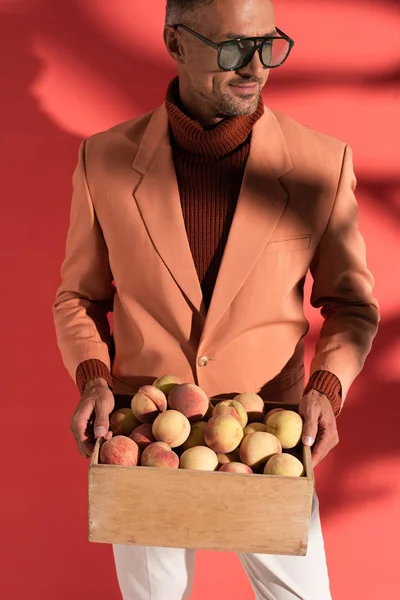 Happy man in sunglasses holding box with sweet peaches on red with shadows — Stock Photo