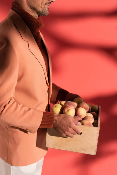 Vue recadrée de l'homme heureux dans le blazer tenant boîte avec des pêches douces sur rouge avec des ombres — Photo de stock