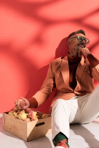 Happy man in blazer and sunglasses sitting near box with sweet peaches on red with shadows — Stock Photo