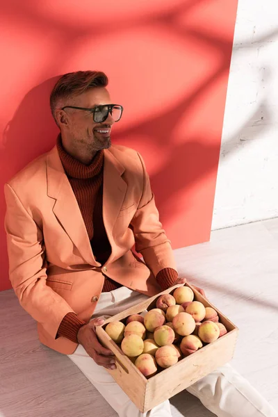 Homme heureux en blazer et lunettes de soleil assis avec des pêches douces en boîte sur rouge et blanc avec des ombres — Photo de stock