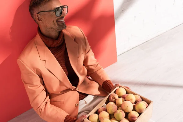 Cheerful man in blazer and sunglasses sitting with sweet peaches in box on red and white with shadows — Stock Photo