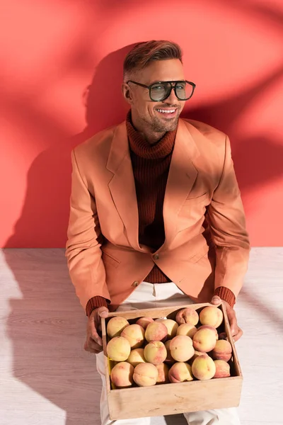 Happy man in blazer sitting with sweet peaches in box on red and white with shadows — Stock Photo