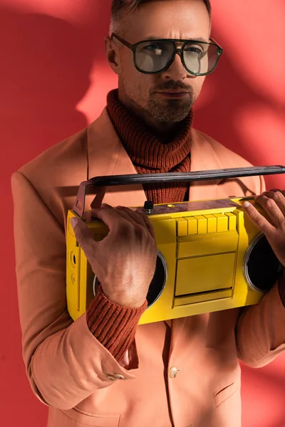 Homme élégant dans des lunettes de soleil tenant boombox rétro sur rouge avec des ombres — Photo de stock