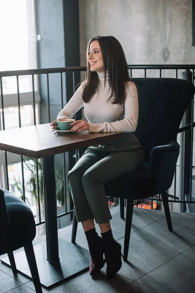 Улыбающаяся молодая женщина, сидящая за деревянным столом с капучино на балконе в кафе — стоковое фото