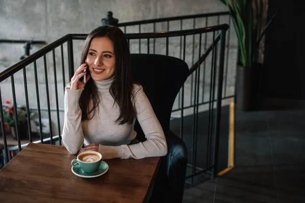 Smiling young woman sitting at wooden table with cappuccino and talking on smartphone on balcony in coffee shop — Stock Photo