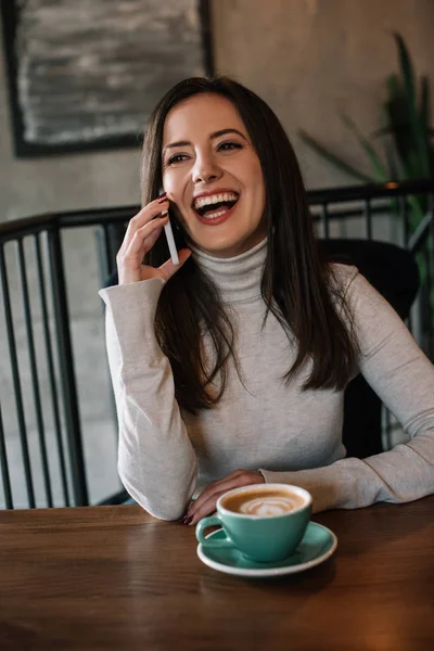 Happy young woman sitting at wooden table with cappuccino and talking on smartphone on balcony in coffee shop — Stock Photo