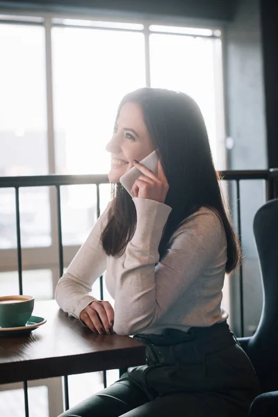 Sorrindo jovem mulher sentada à mesa de madeira com cappuccino e falando no smartphone na varanda no café — Fotografia de Stock