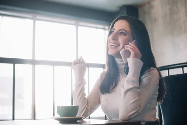 Smiling young woman sitting at wooden table with cappuccino and talking on smartphone on balcony in coffee shop — Stock Photo