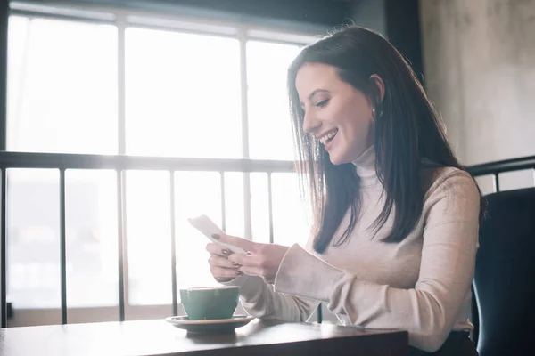 Happy young woman sitting at wooden table with cappuccino and using smartphone on balcony in coffee shop — Stock Photo