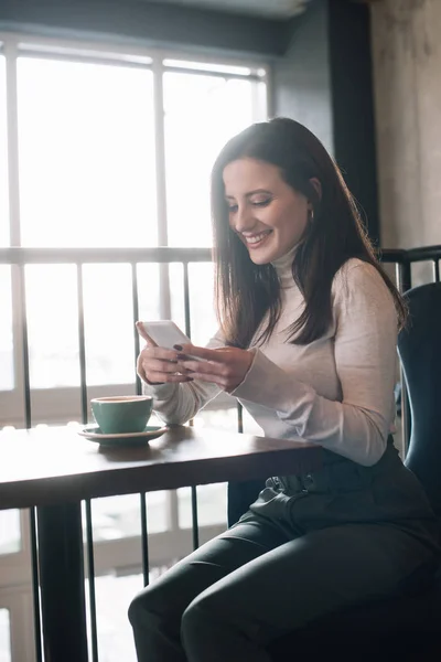 Sorrindo jovem mulher sentada à mesa de madeira com cappuccino e usando smartphone na varanda no café — Fotografia de Stock