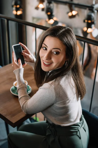 Smiling young woman sitting at wooden table with cappuccino and using smartphone on balcony in coffee shop — Stock Photo