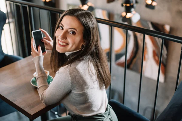 Giovane donna sorridente seduta al tavolo in legno con cappuccino e smartphone sul balcone in caffetteria — Foto stock