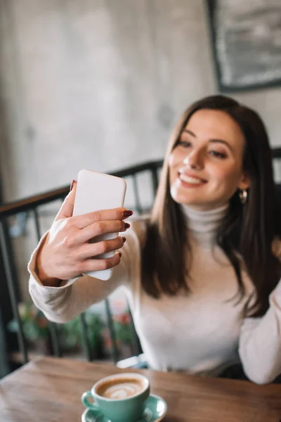 Selective focus of smiling young woman sitting at wooden table with cappuccino and taking selfie on smartphone on balcony in coffee shop — Stock Photo