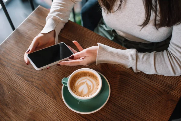 Ausgeschnittene Ansicht einer jungen Frau, die an einem Holztisch mit Cappuccino sitzt und ihr Smartphone mit leerem Bildschirm auf dem Balkon eines Cafés hält — Stockfoto