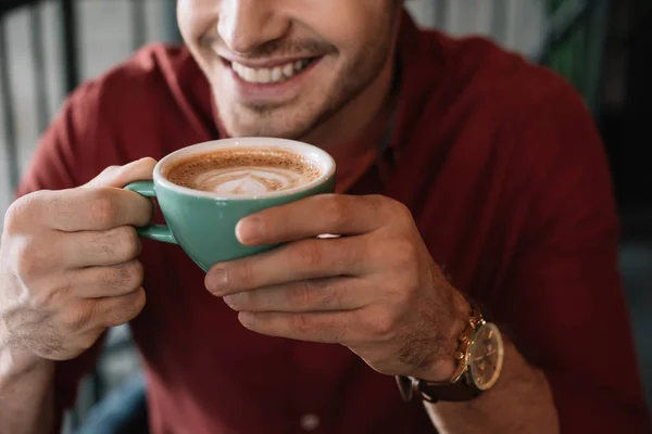 Cropped view of smiling young man with cup of tasty cappuccino in coffee shop — Stock Photo