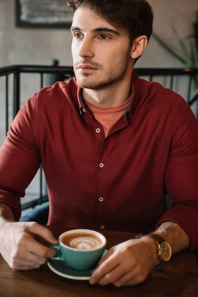 Serous young man sitting at wooden table with cappuccino in coffee shop — Stock Photo