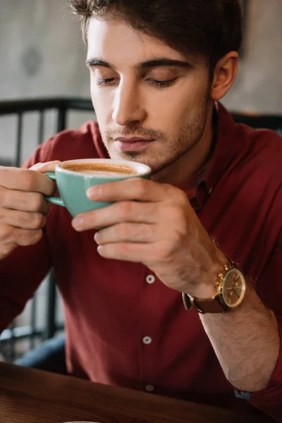Dreamy sad young man sitting at wooden table with cappuccino in coffee shop — Stock Photo