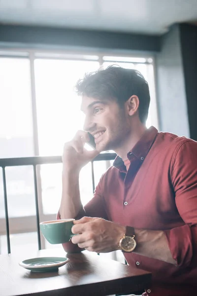 Smiling young man sitting at wooden table with cappuccino and talking on smartphone on balcony in coffee shop — Stock Photo