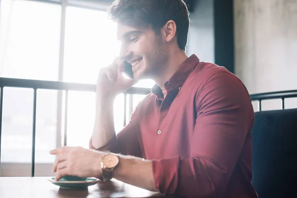 Smiling young man sitting at wooden table with cappuccino and talking on smartphone on balcony in coffee shop — Stock Photo