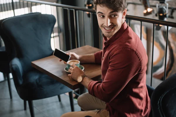 Lächelnder junger Mann sitzt mit Cappuccino am Holztisch und hält Smartphone auf Balkon im Café — Stockfoto