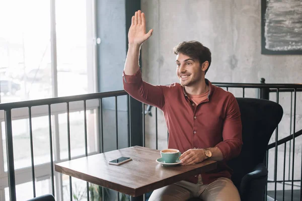 Jovem sorridente sentado à mesa de madeira com cappuccino e smartphone e acenando a mão na varanda no café — Fotografia de Stock