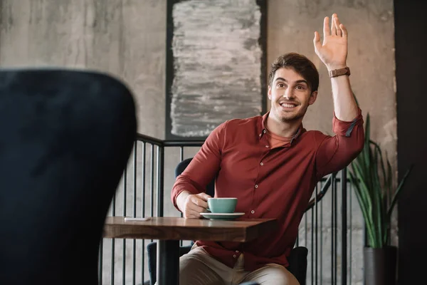 Smiling young man sitting at wooden table with cappuccino and waving hand on balcony in coffee shop — Stock Photo