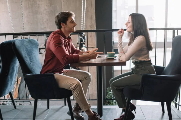 Vista lateral de la alegre pareja hablando mientras bebe café en el balcón en la cafetería - foto de stock