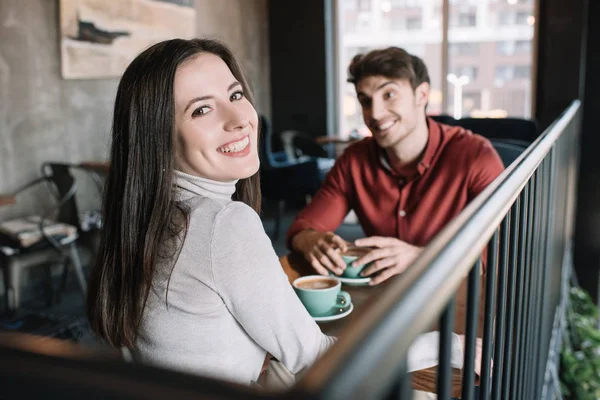 Enfoque selectivo de la chica sonriente mirando a la cámara mientras bebe café con el hombre en el balcón en la cafetería - foto de stock