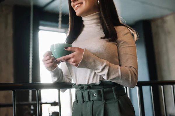 Vista recortada de la mujer sonriente de pie en el balcón con capuchino en la cafetería - foto de stock